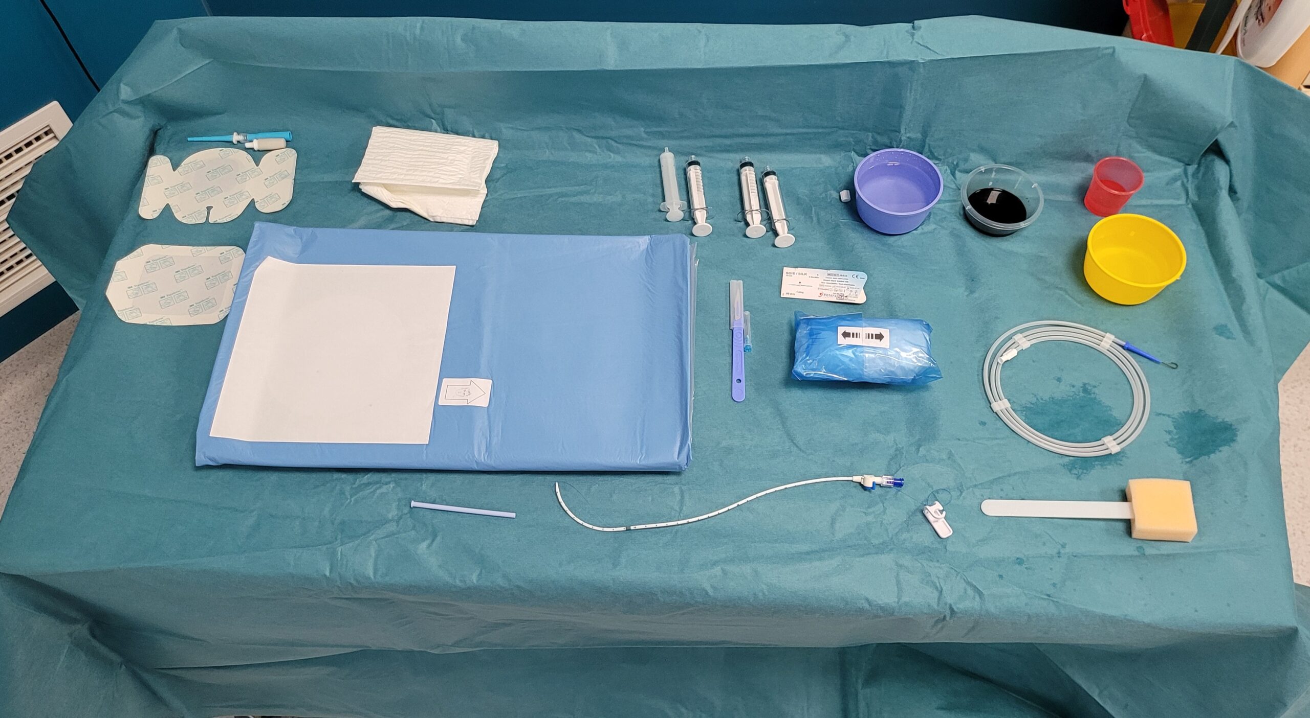 La table d’examen est composée d’un champ de table sur lequel est posé un champ fenêtré qui couvre tout le patient jusqu’au pied, et de cupules qui vont recevoir du sérum physiologique, un antiseptique identique à la détersion, du produit de contraste urinaire et de la lidocaïne. Sur cette table d’examen il y a aussi des compresses, des seringues, une aiguille sous cutané, un scalpel, un guide métallique en J de 150 cm et 0,035 inch de diamètre, et un fil de suture non résorbable type soie si la sonde est fixée à la peau.